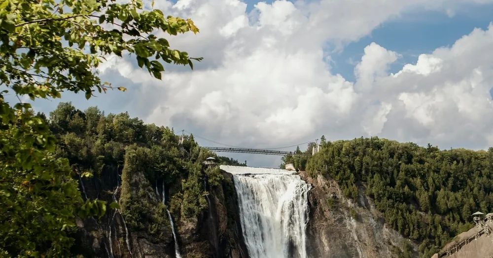 Montmorency Falls framed by green forest under a cloudy sky, with the suspension bridge above the crest
