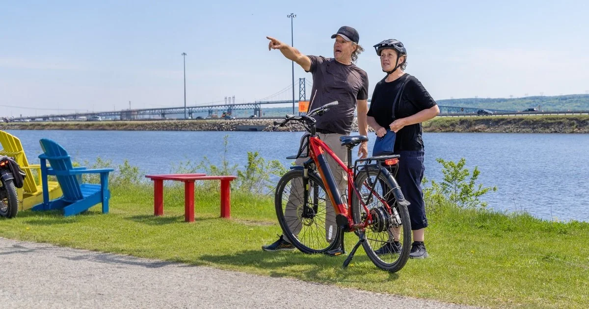 Two cyclists with electric bikes on a grassy riverside path by the St. Lawrence